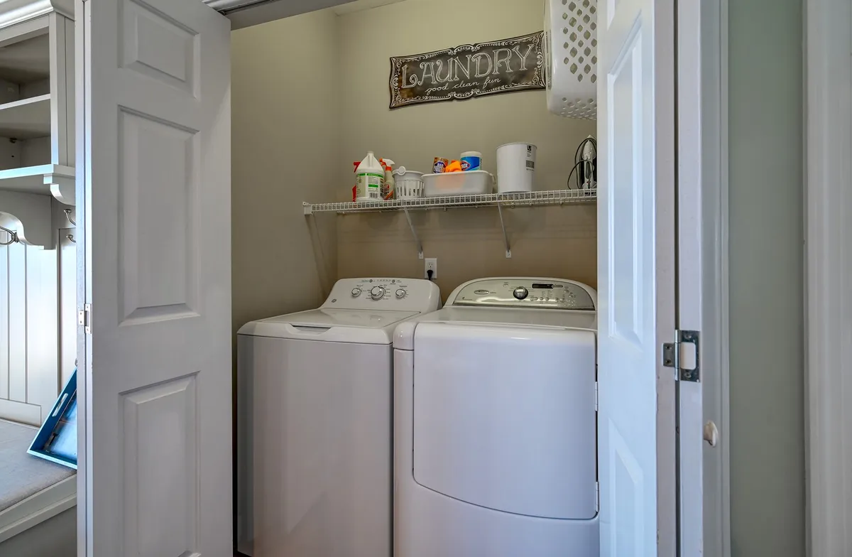 A practical laundry area featuring a washing machine, dryer, a 'laundry' sign, and a shelf with cleaning supplies. The space is tidy, well-organized, and conveniently located for handling post-beach laundry needs.