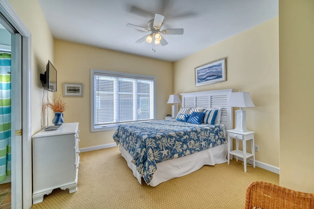 A sunlit bedroom with a bed adorned in blue floral bedding, large window streaming in natural light, white nightstands with lamps, and decorative wall art. The atmosphere is calm, serene, and perfect for unwinding.