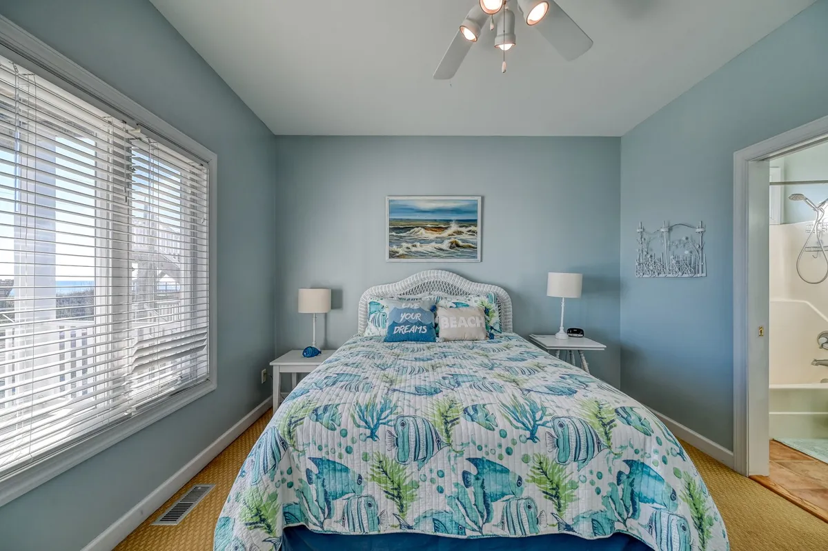 A serene bedroom featuring a bed with ocean-themed bedding, framed beach painting above the headboard, bedside lamps, and decorative pillows. The large window lets in abundant natural light, creating a calm, inviting atmosphere perfect for relaxation.