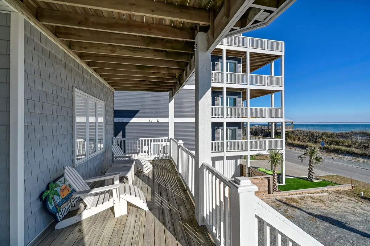 A serene exterior porch scene with adirondack chairs arranged to take in sweeping beach and ocean views, framed by palm trees and a wooden railing. The space exudes relaxation, perfect for family conversations, quiet mornings, or evening gatherings overlooking the Atlantic.