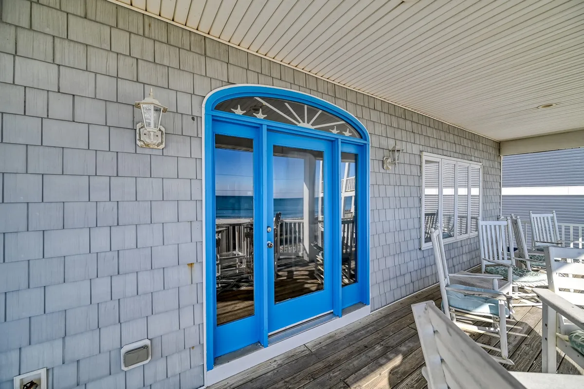A charming oceanfront porch with white rocking chairs, blue double doors, and a wooden deck overlooking the waves. The scene invites guests to relax, sway in a rocker, or enjoy a meal outdoors as the sun sets.
