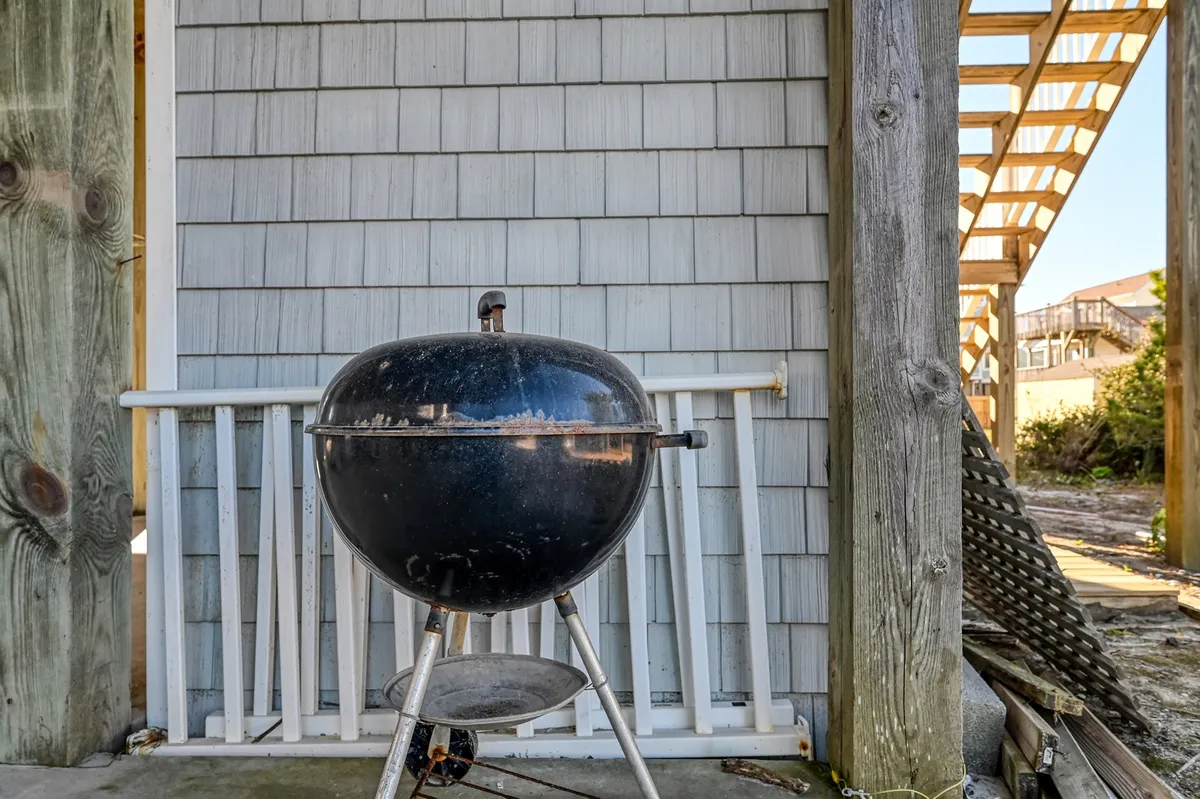 A casual exterior view featuring a wooden structure with shuttered railings, a grill set up for barbecuing, and ample porch space for outdoor gatherings. The scene suggests a relaxed, communal atmosphere ideal for group activities and shared meals.