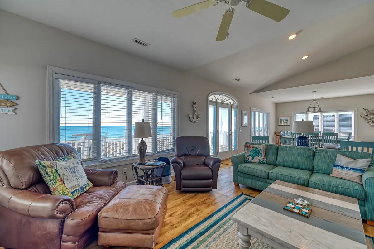 A bright, open living room with large windows framing stunning ocean views. Comfortable sofas arranged for conversation and relaxation fill the space, surrounded by decorative throw pillows and a coffee table with coastal accents. The entire scene is flooded with natural light, creating a cozy yet spacious atmosphere.