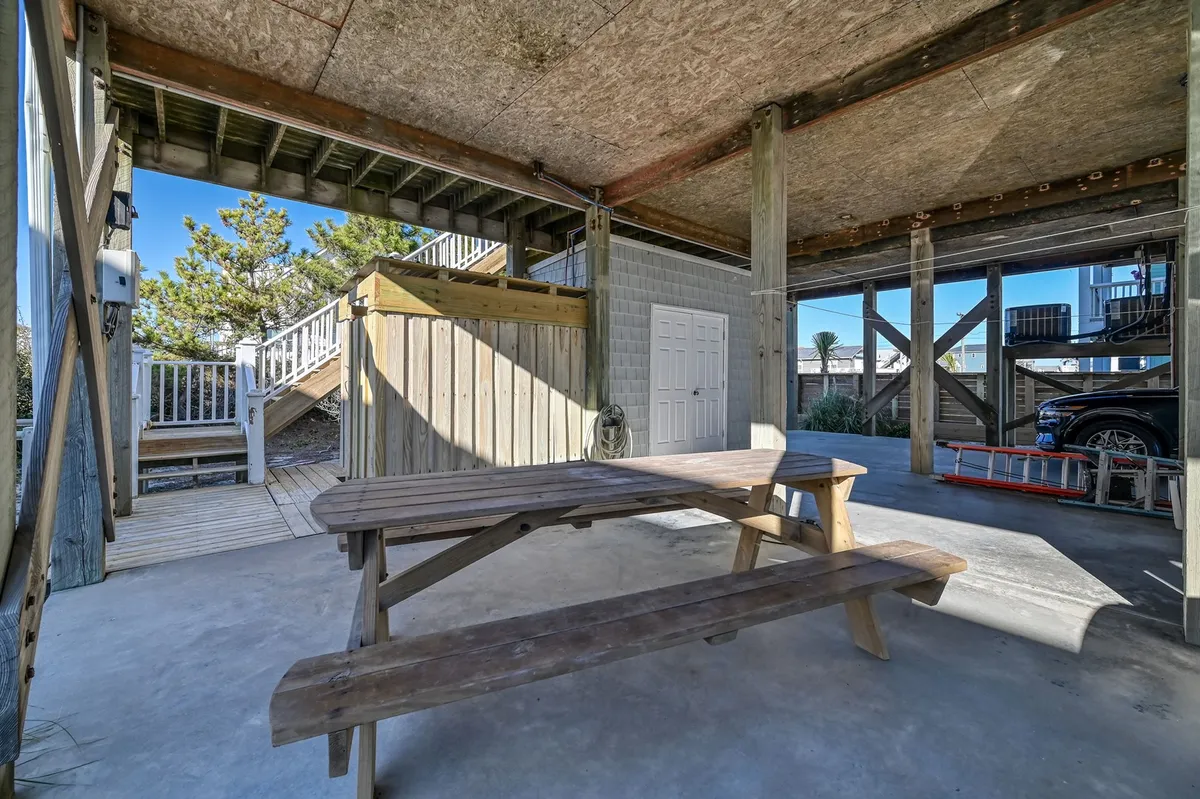 A spacious exterior area featuring a sturdy wooden picnic table beneath the shade of wooden beams, with a staircase leading up. The setting is ideal for family barbecues, game nights, and sunset dinners after a day spent on the beach or pier.