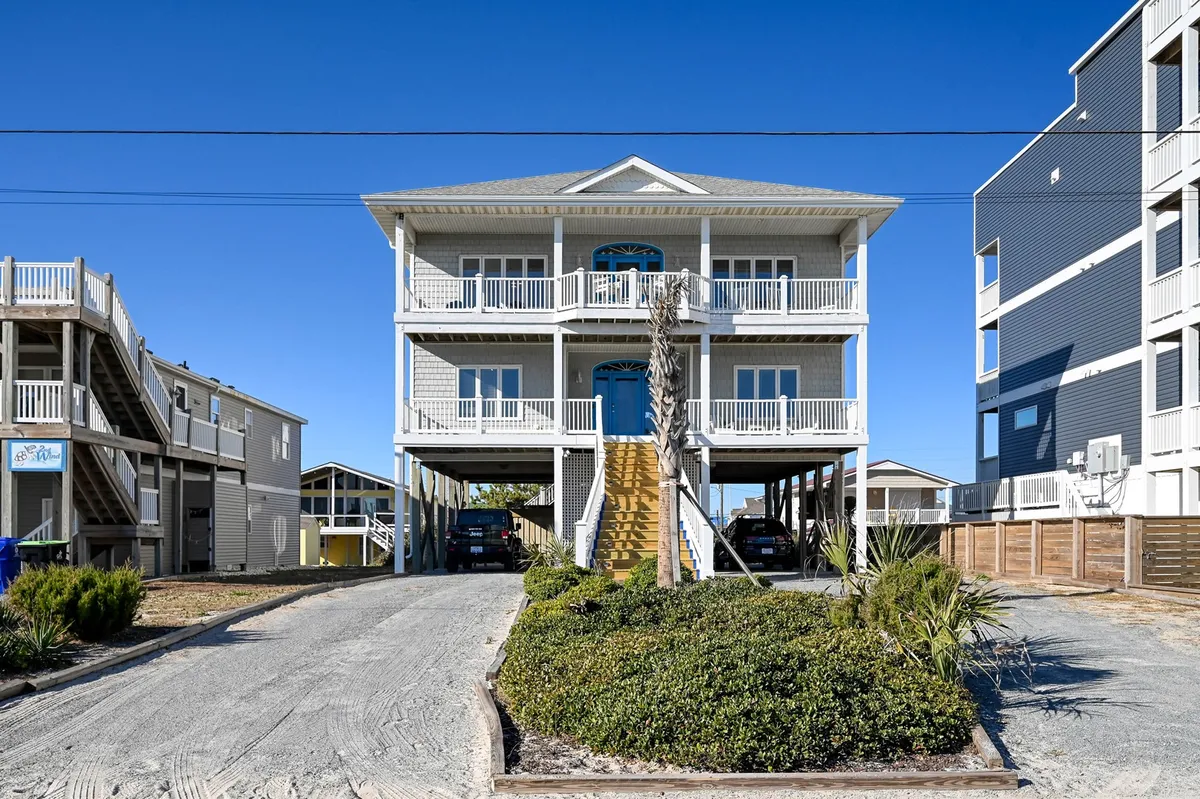 This exterior shot displays a welcoming bright blue front door, spacious balconies, and a driveway leading up to the house. The area under the balcony offers covered parking, visible in the foreground. The inviting setting suggests easy arrivals and sheltered access for guests and their pets.