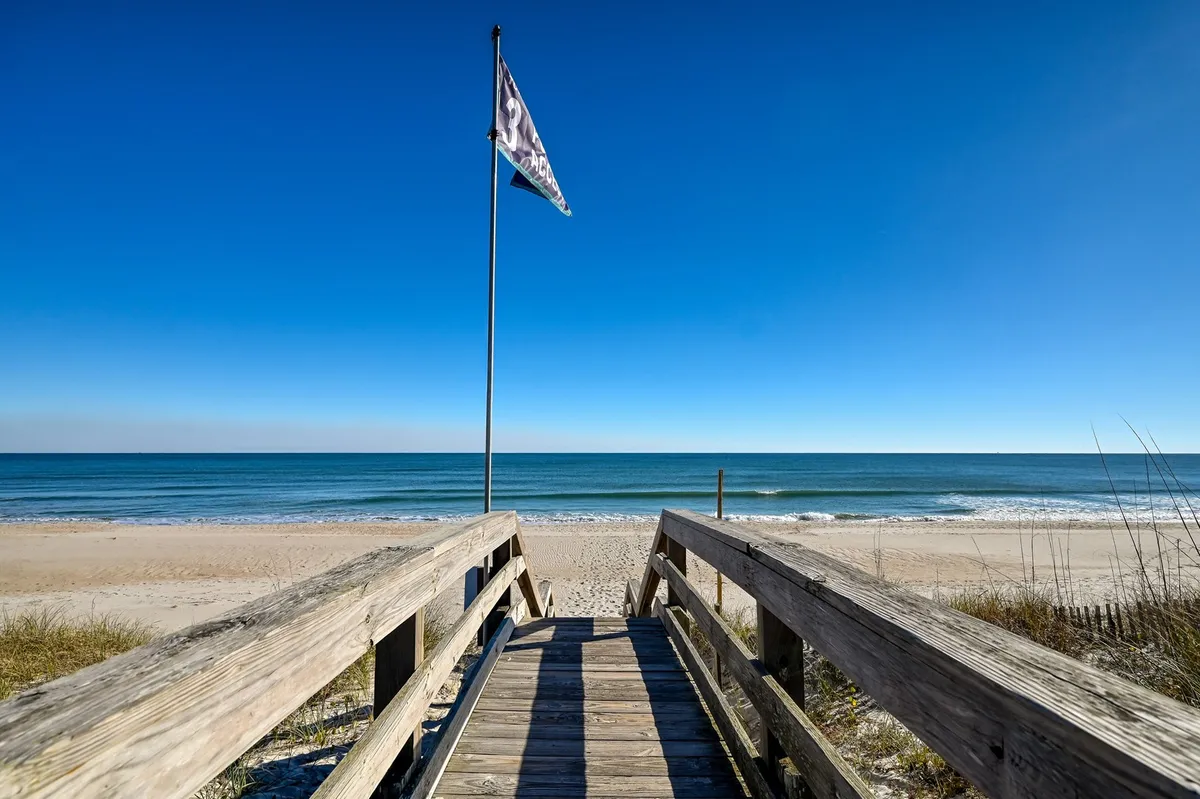 A wooden walkway leads directly to the sandy beach, with the blue ocean shimmering in the background and a small flag waving in the coastal breeze. The scene shows a clear connection between the home and the oceanfront, inviting families to stroll onto the sand for sunbathing or play.