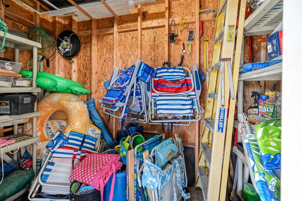 An interior space filled with stacked beach chairs, colorful pool floats, storage bins, and a ladder—visibly ready for a large group’s beach day or organized vacation prep. The scene feels playful and functional, suggesting flexibility and DIY options.