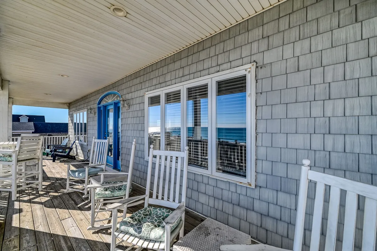 An inviting exterior porch with rocking chairs facing an ocean view and a blue door, framed by evening light. The scene evokes peaceful relaxation, group conversations, and the scenic setting of a beachfront escape.