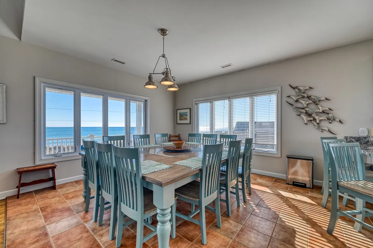 A grand solid wood dining table sits in front of wide windows showcasing the ocean, comfortably seating ten guests. Decorative fish art on the wall adds a playful, coastal touch. The arrangement is perfect for family dinners, games, or morning breakfasts with sunlight streaming in.