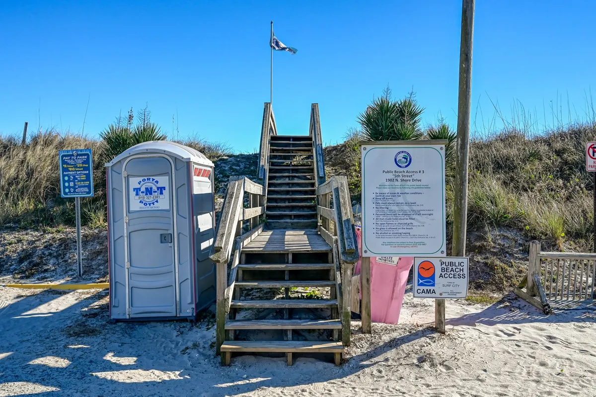 A set of stairs leads directly to the sandy beach, with a clearly marked beach access sign and a portable restroom nearby. The atmosphere is open and welcoming, suggesting the beach is only moments away from the property.