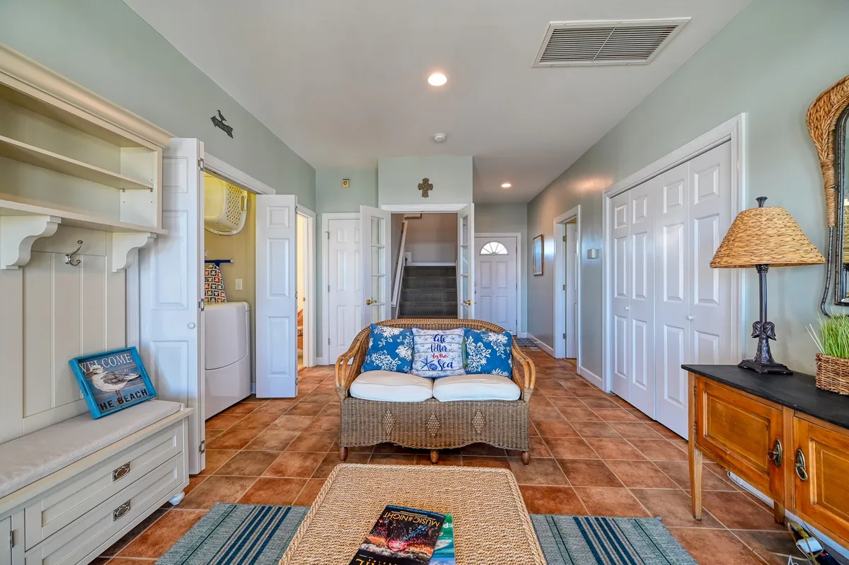 A view of a welcoming entryway with a woven couch, coffee table, decorative wall art, lamp, and an entrance door. The space feels inviting and relaxed, suggesting it is the first impression guests receive upon arrival. Decorative elements and comfortable seating create an immediate sense of home and relaxation.