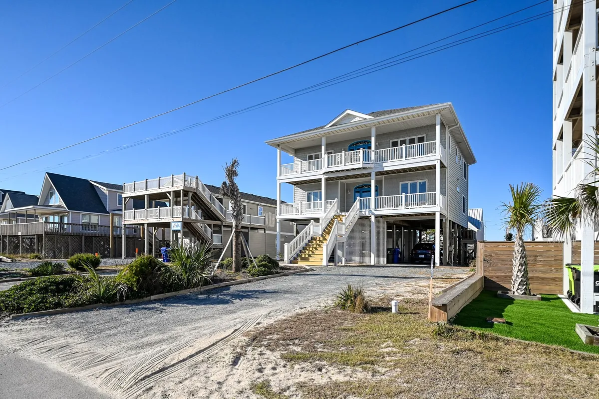 A cheerful two-story house surrounded by palm trees, with a visible staircase leading up to a welcoming porch. The spacious driveway and inviting balconies suggest ample room for large group arrivals and family gatherings. The overall scene exudes a sense of relaxed, multigenerational togetherness.