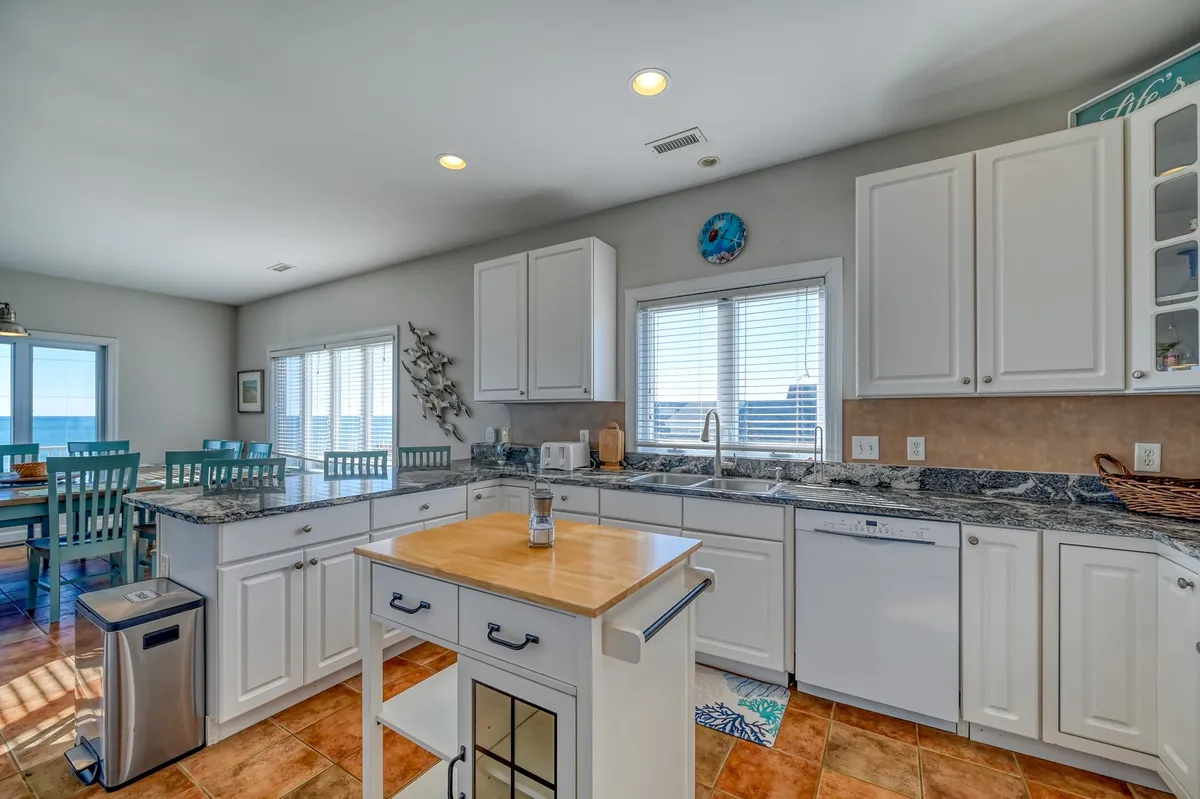 A bright, fully equipped kitchen with a central island, windows offering views of the outdoors, and stylish blue dining chairs. Modern appliances and granite countertops make meal prep a joy while bar seating encourages guests to hang out and socialize.